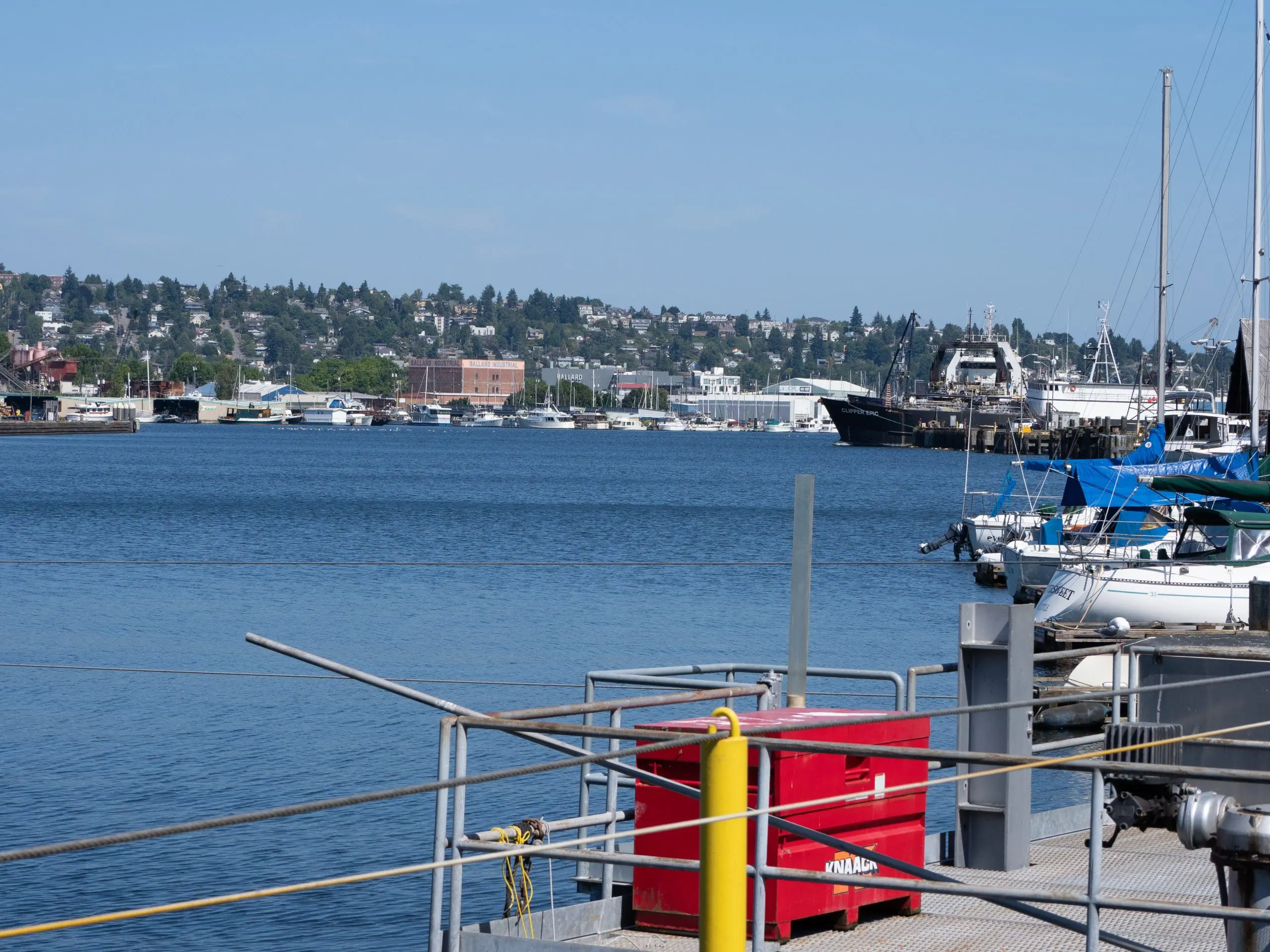 Image is a view across the Lake Washington Ship Canal from a platform with metal railings and a red metal box, looking over several docked sailboats and fishing vessels towards trees, buildings, and boats on the opposite shoreline.