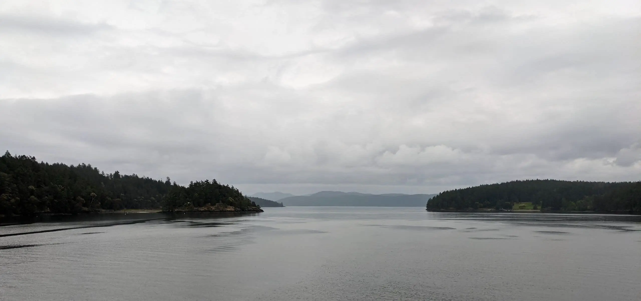 Photo of a wide, grey body of water narrowing to a channel in the center between two green tree-covered land masses. Behind them in the distance are more hills and a cloudy sky.