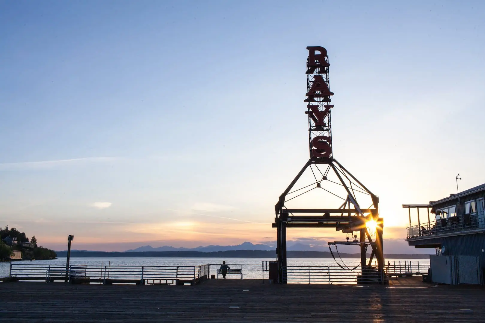 Photo of a large vertical sign reading "RAYS", unlit, silhouetted against the Ballard waterfront with the sun setting behind the Olympic mountains.
